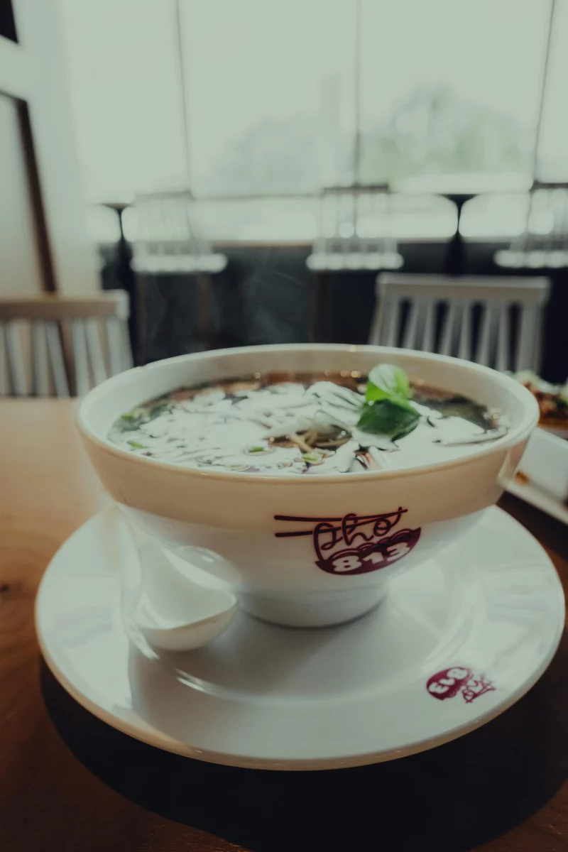 Close-up of a steaming bowl of Vietnamese pho with herbs, captured indoors at a restaurant.