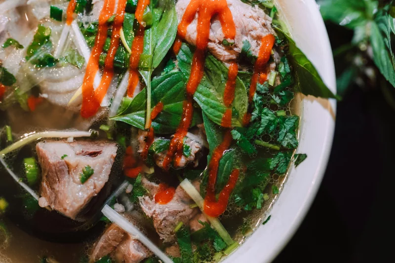 Close-up of Vietnamese pho with fresh herbs, beef, noodles, and Sriracha sauce.