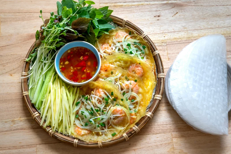 Top view of vibrant Vietnamese Banh Xeo with fresh herbs and a side of chili sauce.