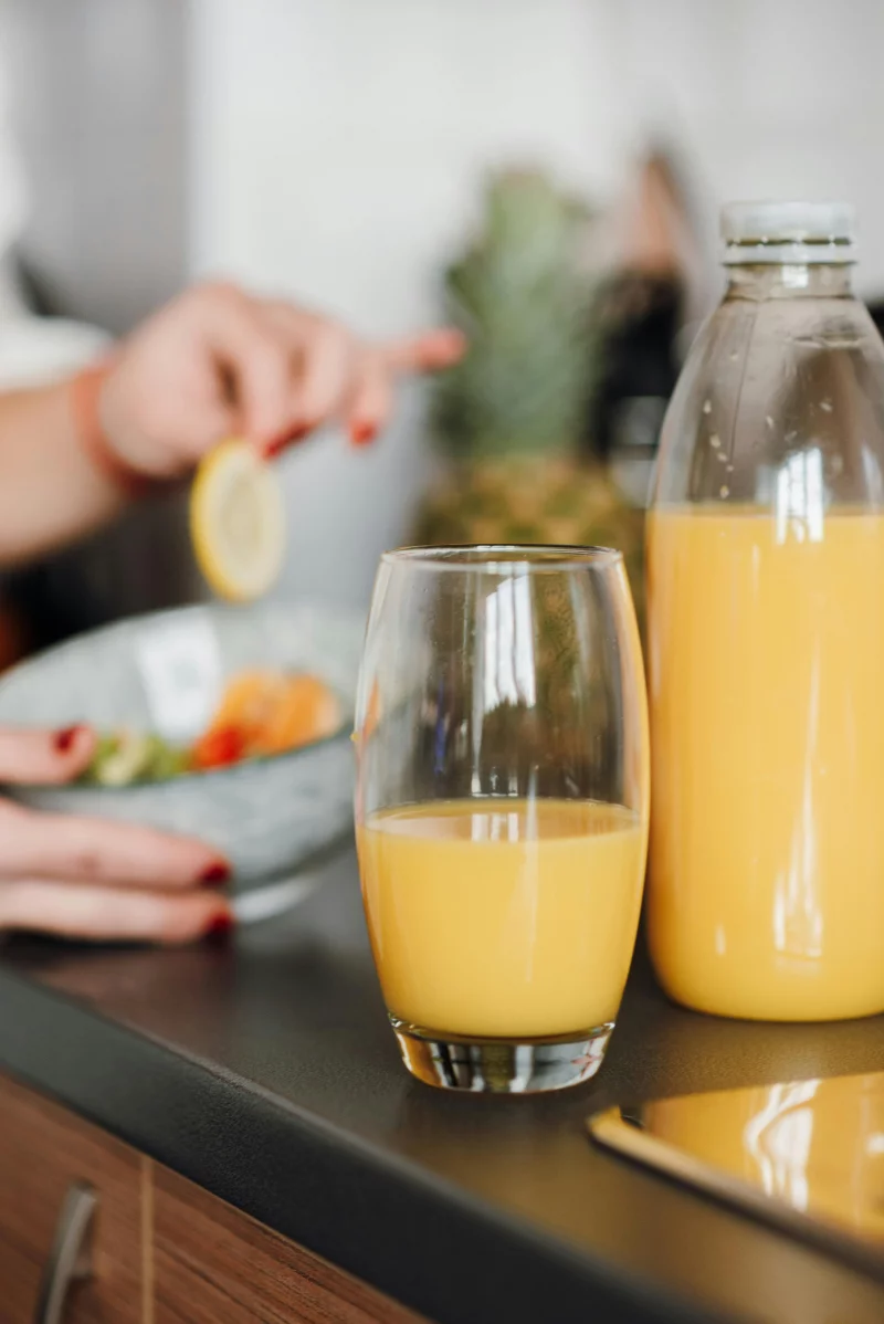 Glass and plastic bottle with natural fruit juice on table near bowl with salad against crop blurred lady having meal