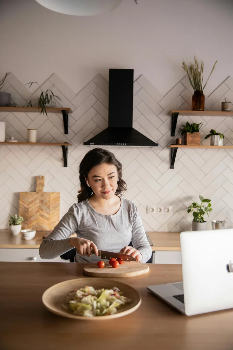 Woman using laptop to follow a recipe while cutting vegetables in a modern kitchen.