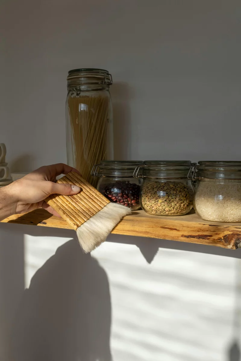 Neatly arranged pantry shelf with jars of grains and pasta, cleaned with a hand-held brush.