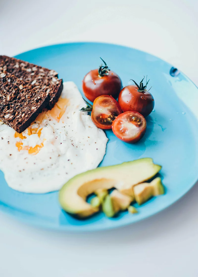 A vibrant and nutritious breakfast plate featuring avocado, sunny side up egg, whole grain toast, and ripe tomatoes.