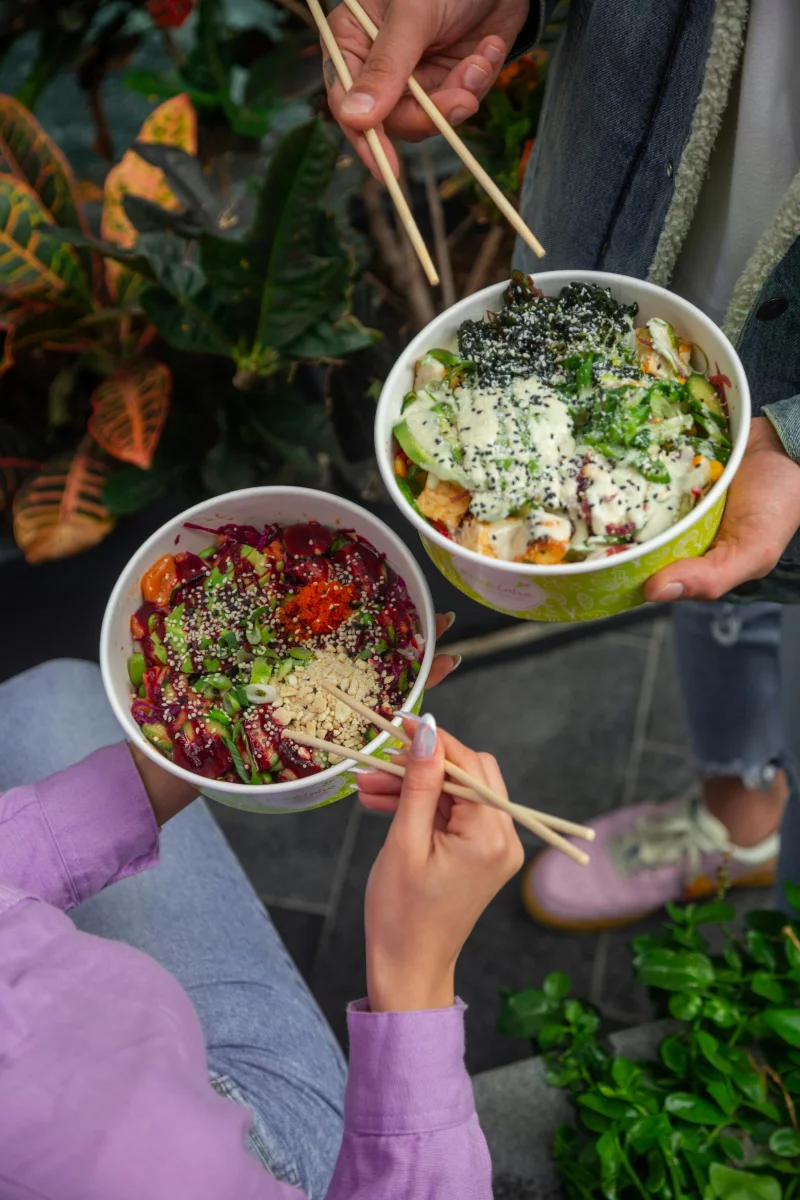 Two people holding colorful salad bowls with chopsticks, surrounded by greenery.