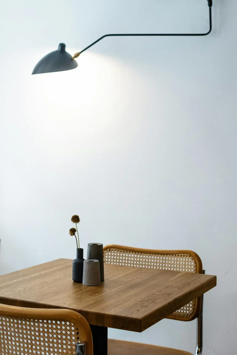 Square shaped wooden table with decorative flower and wicker chairs back and hanging lamp turned on near white wall in flat