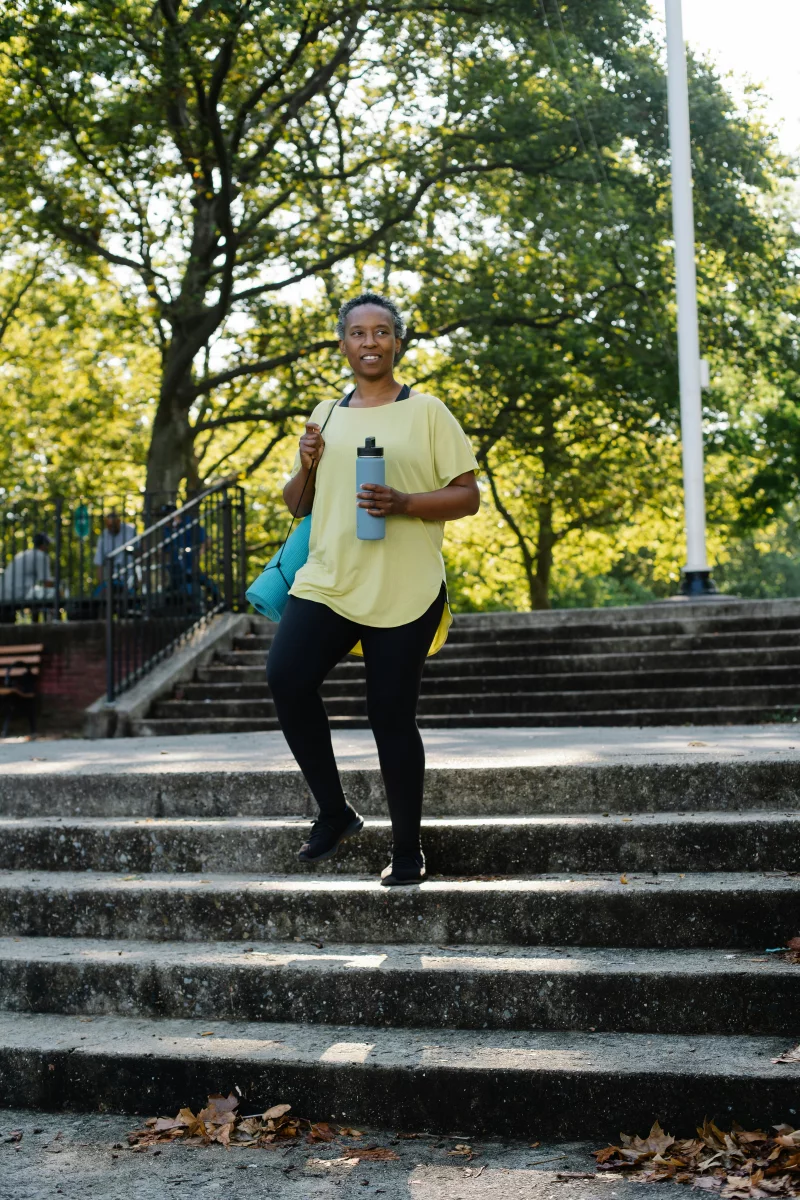 Elderly woman in active wear holding a water bottle standing on park stairs during daytime workout.