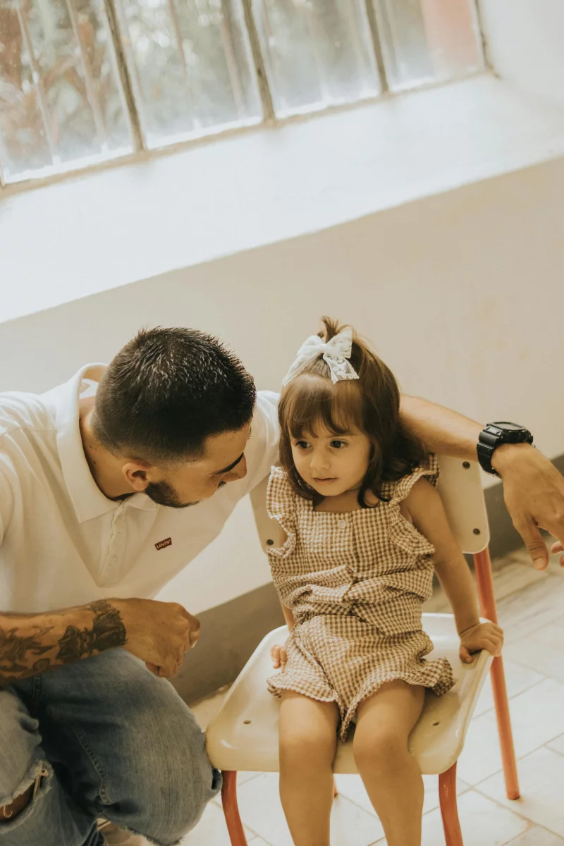 A father crouches beside his daughter in a sunlit room, offering comfort.
