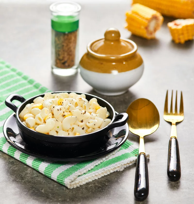 A bowl of creamy corn macaroni with spices and utensils on a rustic tabletop.