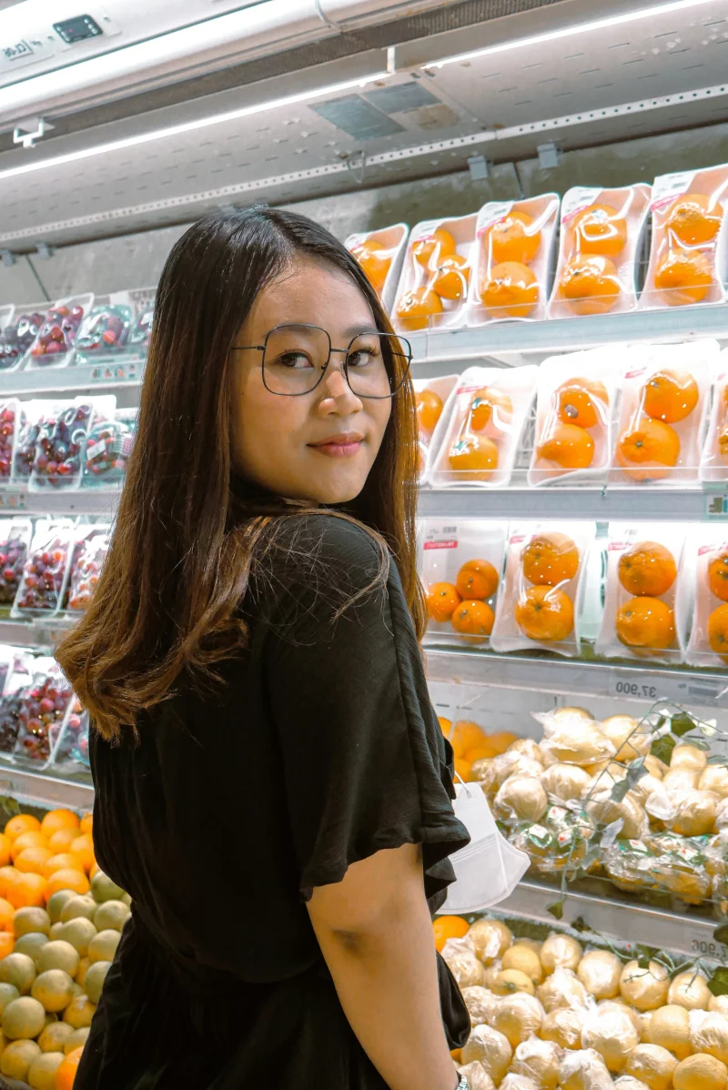 A woman stands in a supermarket produce aisle, looking back at fresh fruits on the shelves.