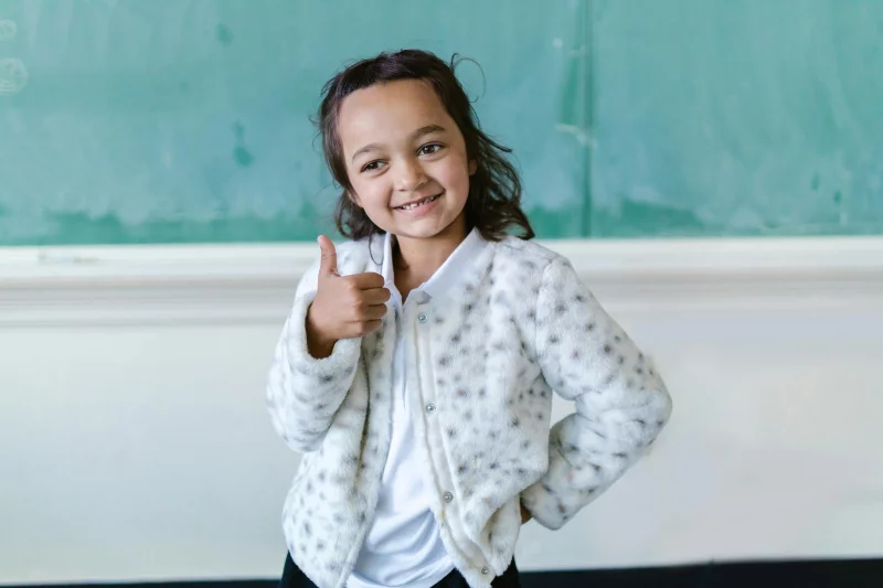 Cheerful young girl giving thumbs up in a classroom setting, conveying positivity.