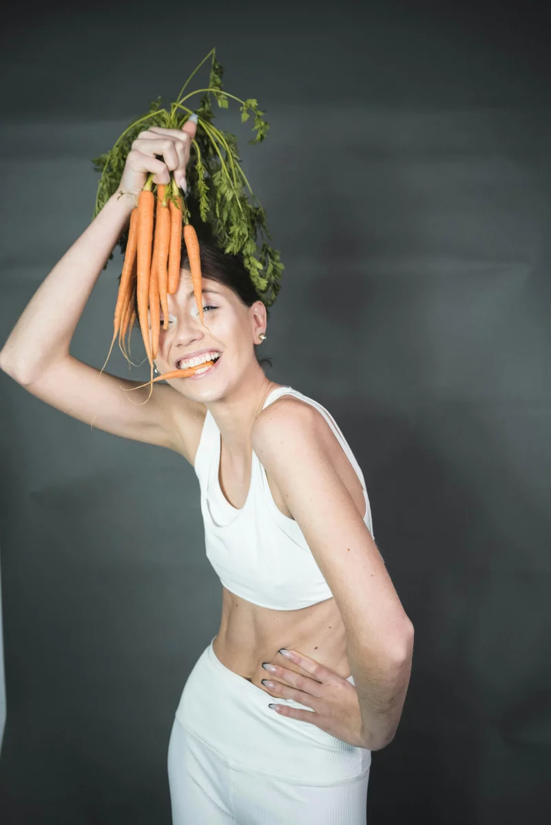 A happy woman in white outfit holding fresh carrots, posing against a gray background.