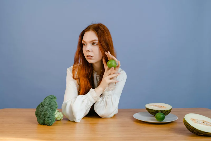 Caucasian woman sitting at table holding lime with broccoli and melon.