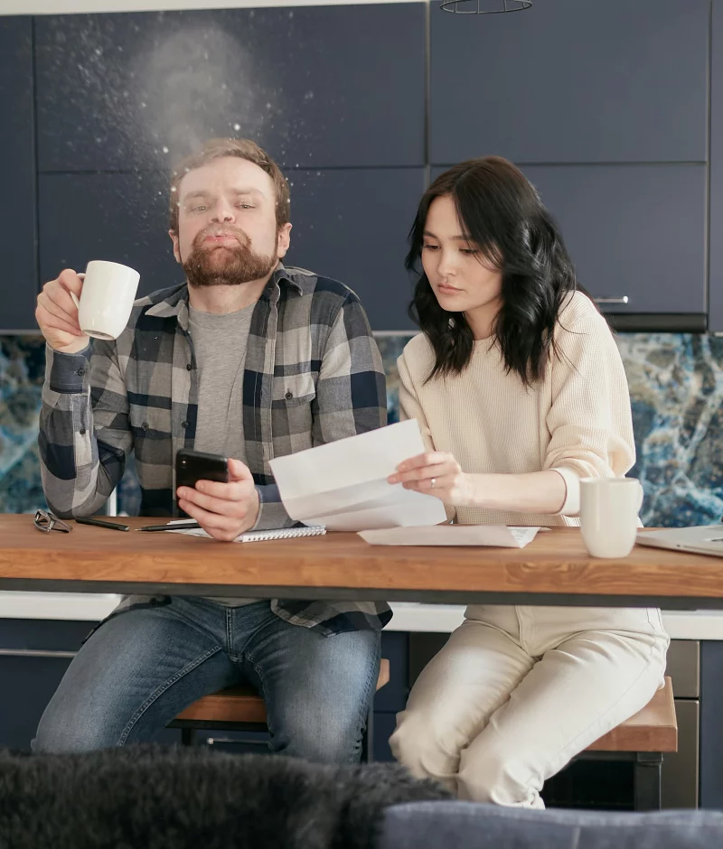A young couple reviews bills and documents in their kitchen, expressing financial stress and concern.