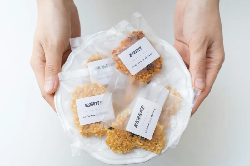Overhead view of hands holding a plate of assorted packaged cookies on a white background.