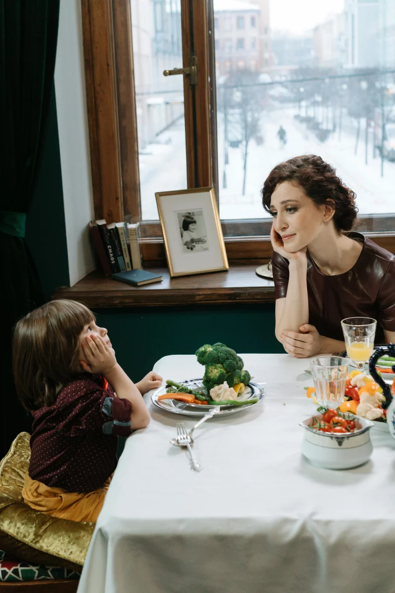 A mother and her child enjoy a meal with vegetables in a cozy dining room.