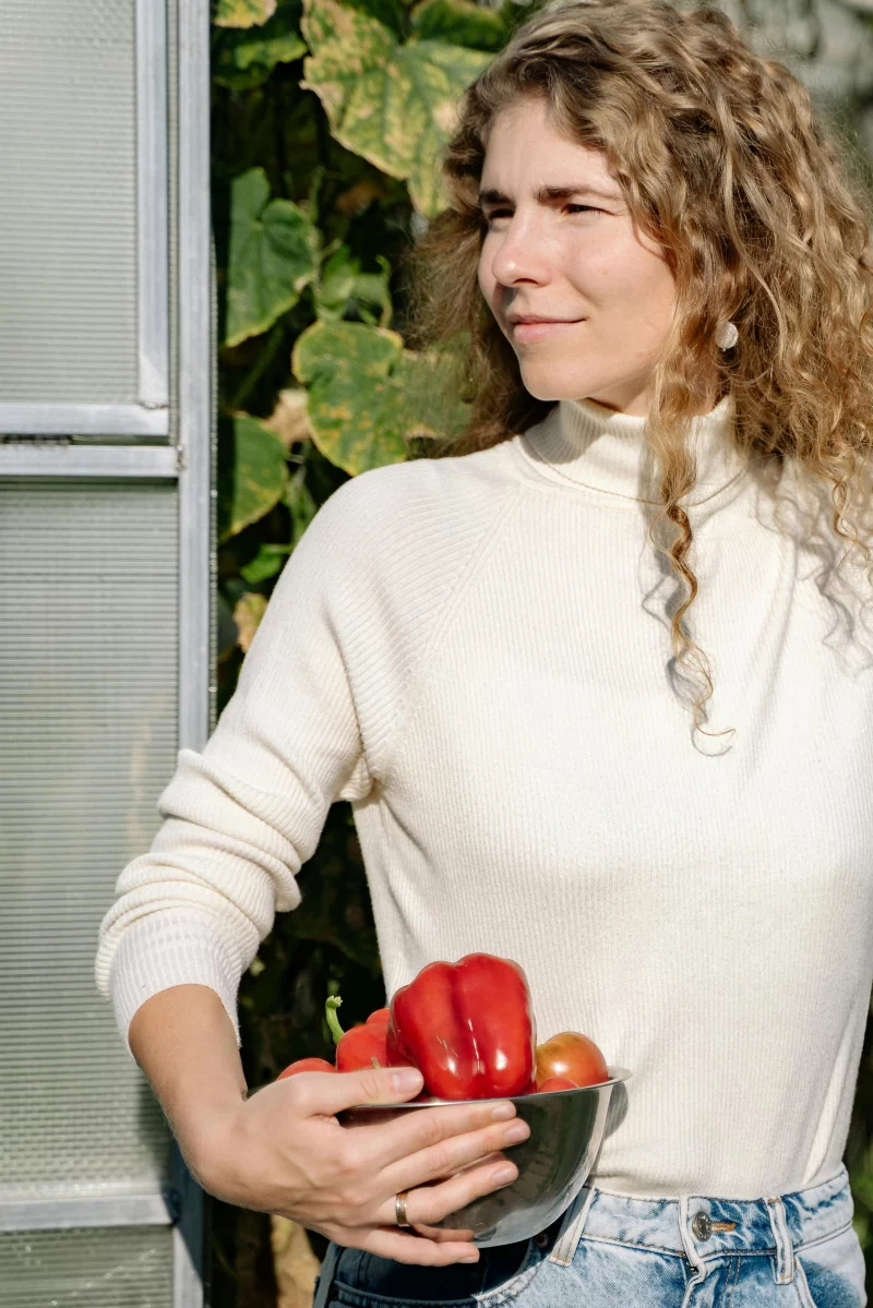A woman in a white sweater holds a bowl of fresh vegetables outdoors.