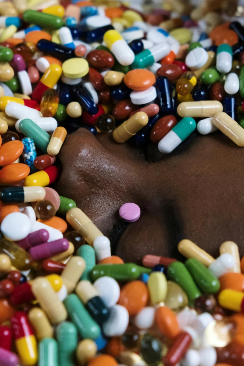 A person's face surrounded by a variety of colorful pills and tablets, symbolizing medication or overconsumption.