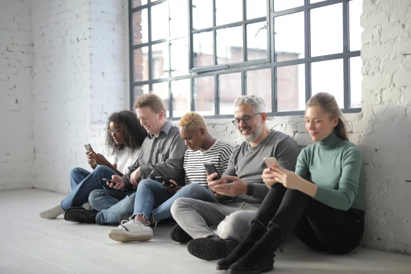 A diverse group of adults seated indoors, engaging with their smartphones in a bright room.