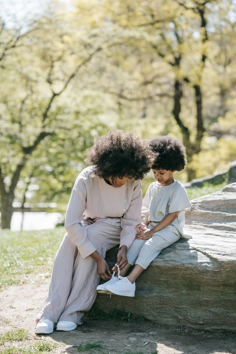 A mother helps her daughter tie shoelaces while sitting on a rock in a park during spring.