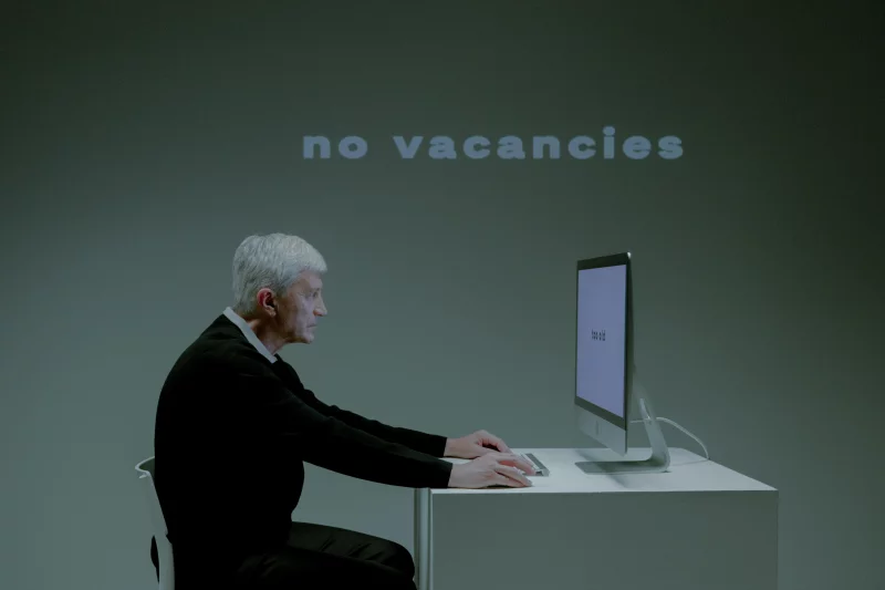 Senior man sitting at a computer with 'no vacancies' displayed, highlighting unemployment issues.