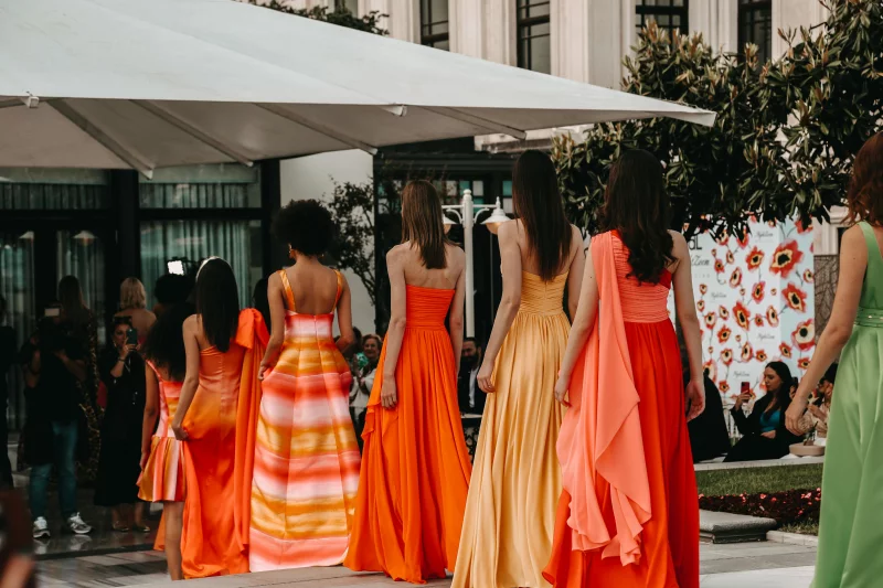 Models in stunning vibrant dresses walk the runway in an outdoor fashion show.