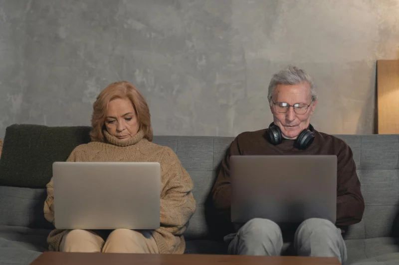 An elderly couple sitting on a sofa at home, each using a laptop. Modern technology engagement.