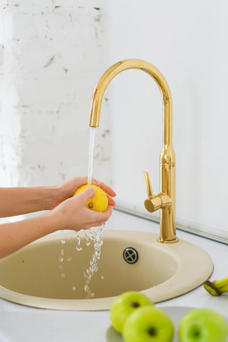 Hands washing a lemon in a sink under running water with green apples nearby.