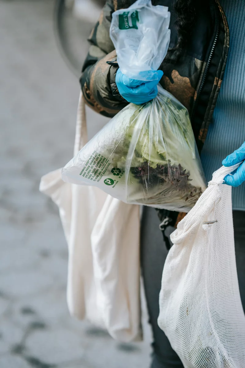 A person wearing gloves shops with eco-friendly bags at an outdoor market. Plastic and mesh bags are visible.