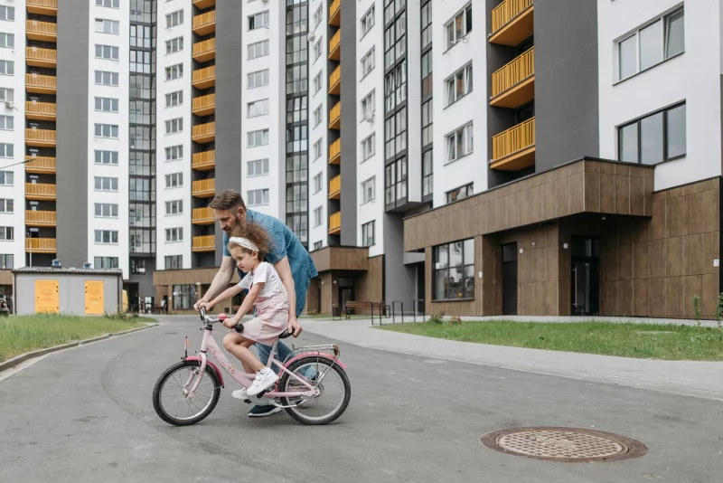 Father guiding daughter on a bike ride outside modern apartments. Bonding and teaching moment.
