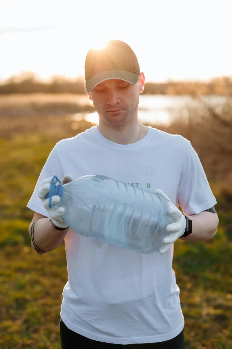 Man in white t-shirt and cap holding an empty plastic bottle outdoors at sunset.
