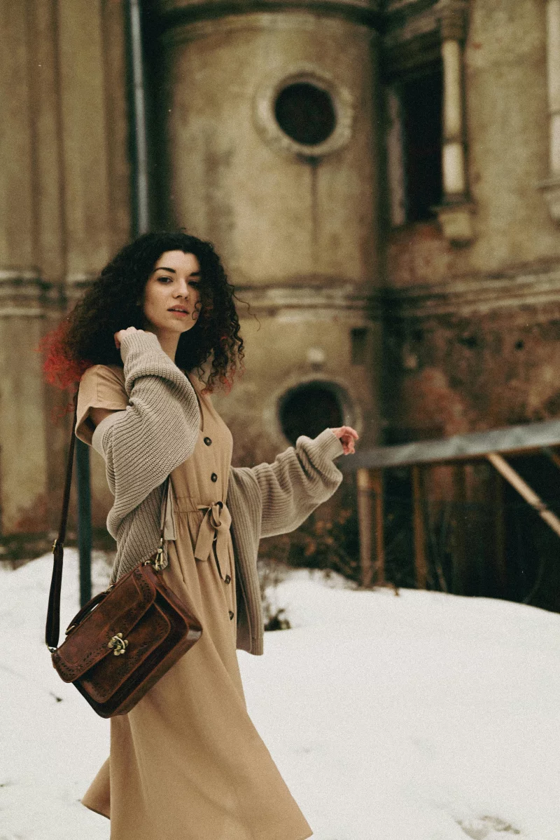 A fashionable woman with curly hair and a handbag poses in a snowy winter setting.