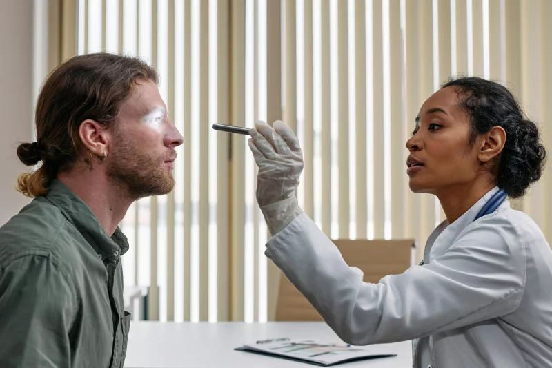 A doctor performs an eye examination on a patient using a flashlight in a clinical setting.