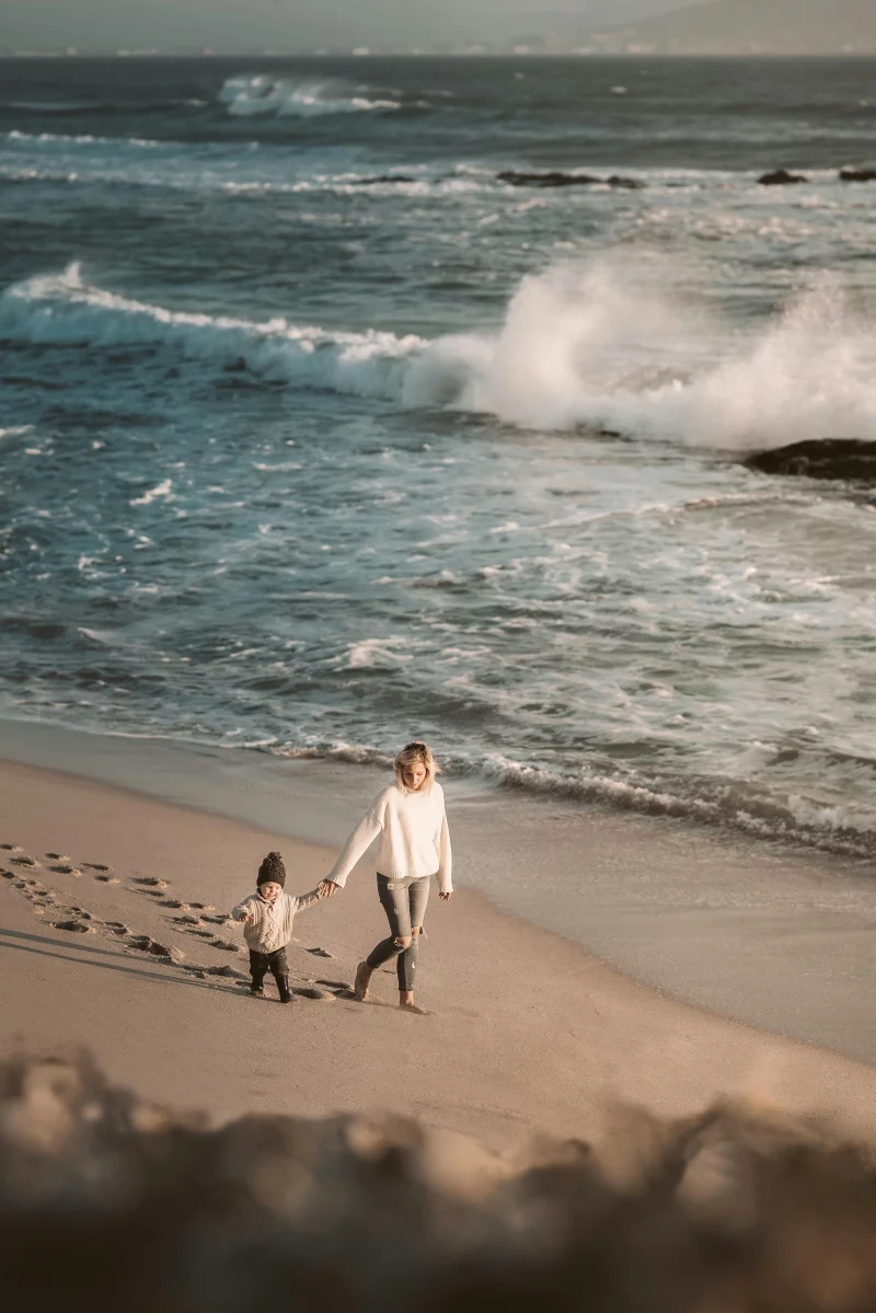 A mother and child walk along the beach with crashing waves in the background, capturing a serene family moment.