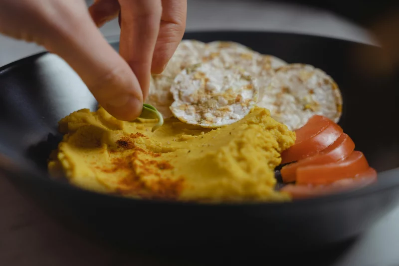 A delightful vegan snack featuring creamy hummus, rice cakes, and fresh tomato slices served in a stylish black bowl.