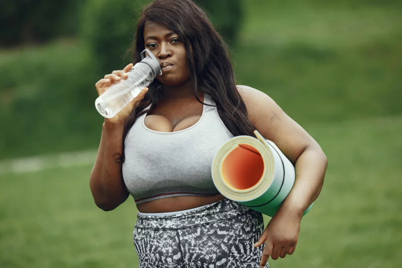 A woman drinks water while holding a yoga mat outdoors, embracing a healthy lifestyle.