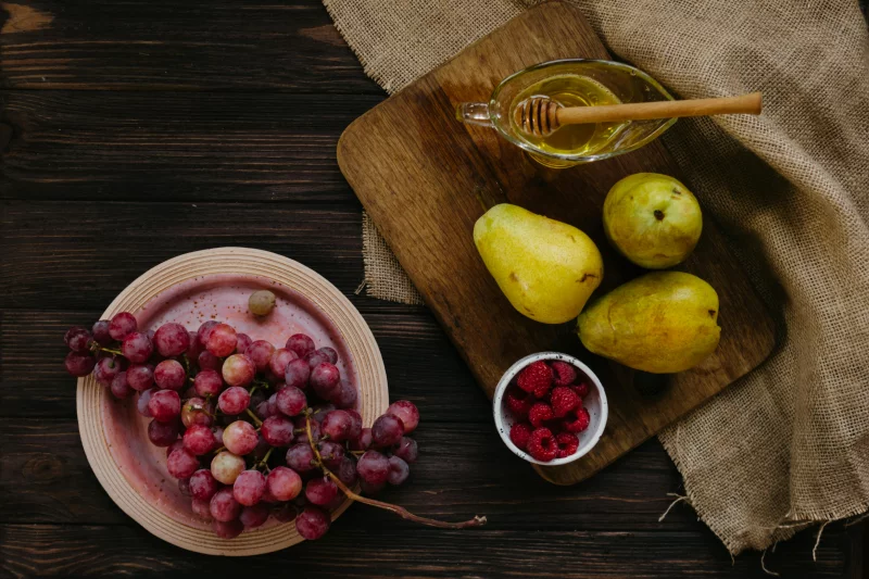 A flat lay of grapes, pears, raspberries, and honey on a wooden board, enhancing healthy eating vibes.