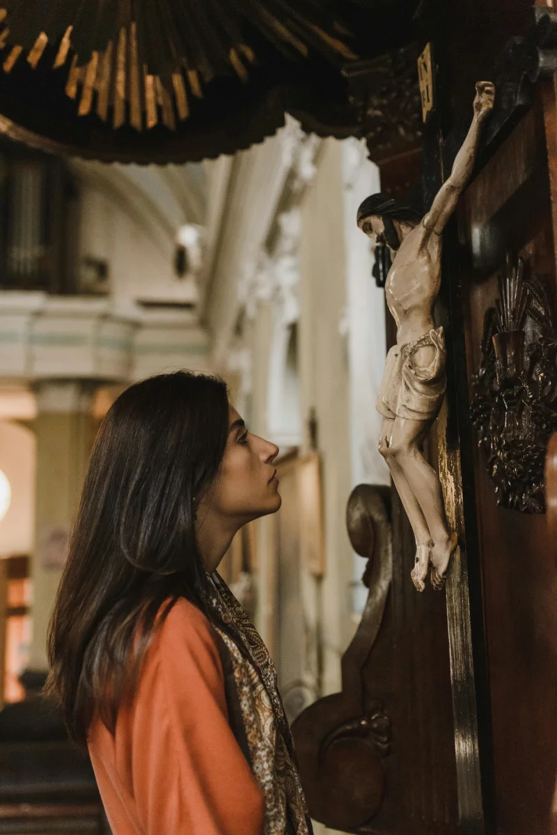 A woman prays beside a detailed sculpture of Jesus in a church interior, capturing a moment of faith.