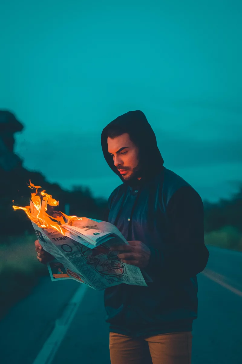 A man in a hoodie reads a burning newspaper on a dimly lit road, creating a dramatic scene.