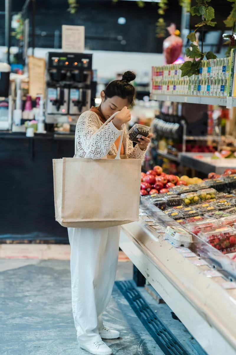 An Asian woman shopping for fresh fruit in a supermarket, holding a tote bag.