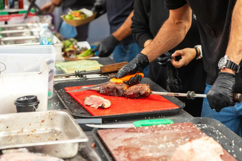 Chefs slicing grilled meat at an outdoor food market with fresh ingredients visible.