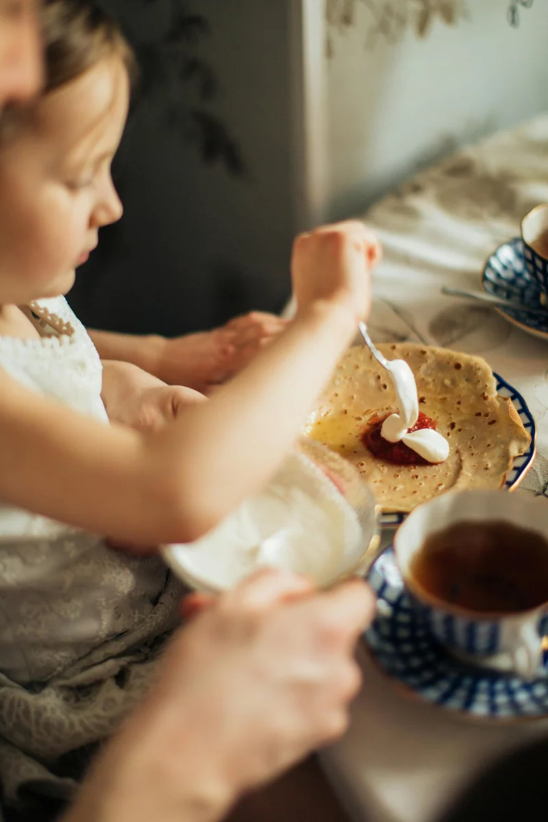 A child and parent enjoy making crepes with cream and jam in a warm, cozy kitchen setting.