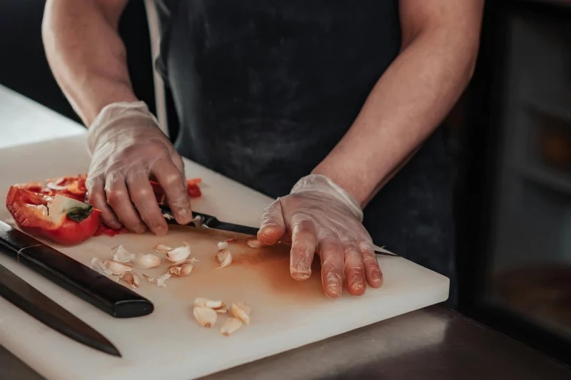 A chef wearing gloves slices garlic and red pepper on a chopping board, showcasing precision and hygiene.