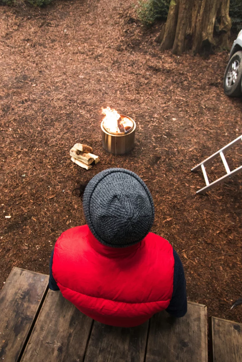 Person in red vest enjoys a campfire on a chilly day in a forest setting.