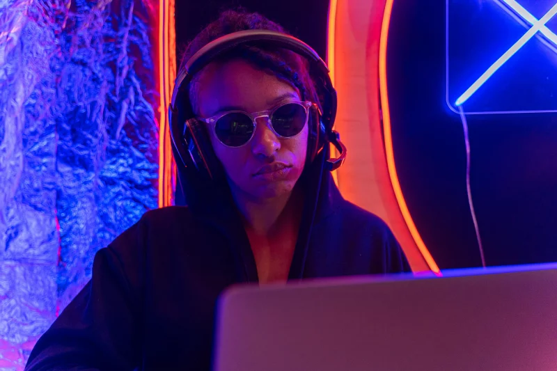 Person in sunglasses and headphones using a laptop in a neon-lit setting.