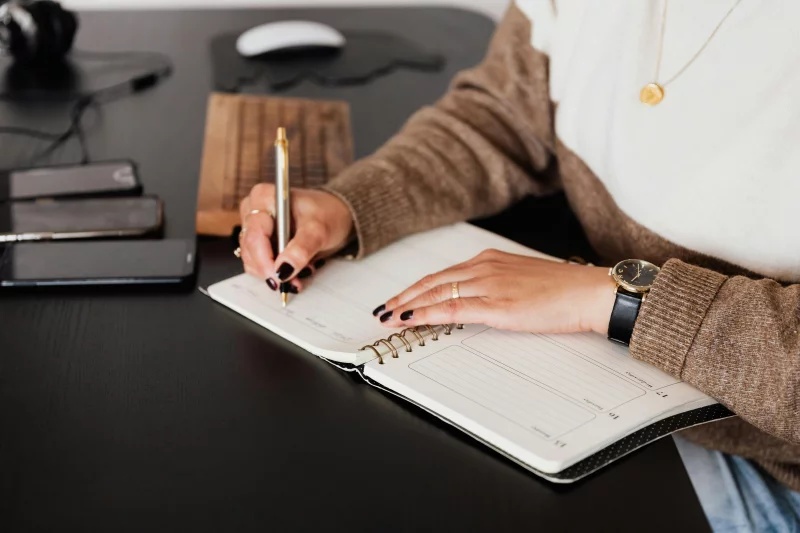 A female freelancer notes plans in a diary at a sleek desk with devices.