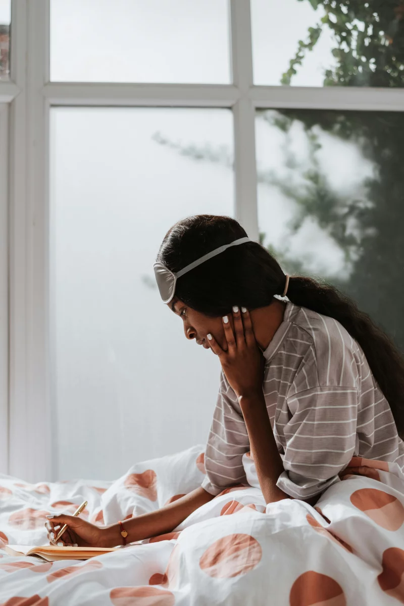 African American woman in pajamas writing in bed by the window, captured in natural light.