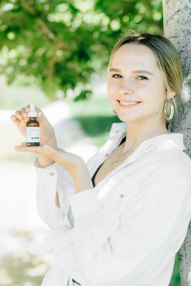 A young woman smiles while holding a CBD oil bottle outdoors in a sunny park.
