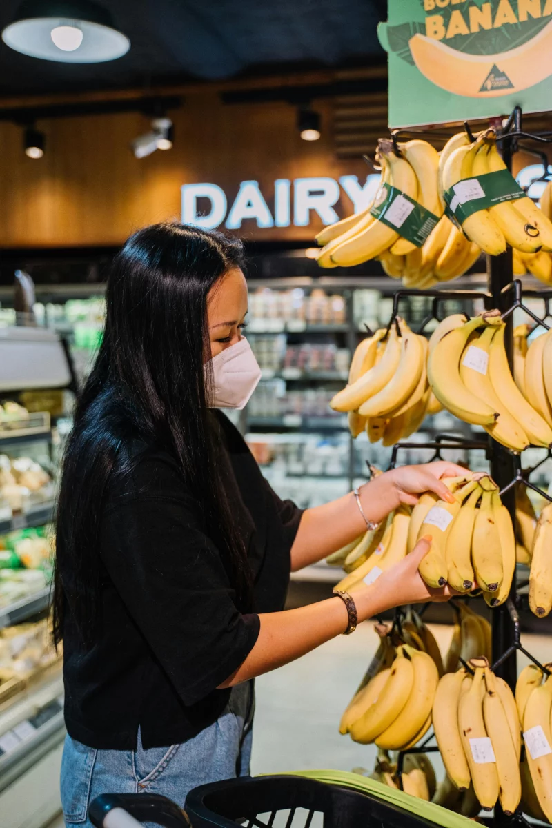 A woman wearing a mask selects ripe bananas in a grocery store fruit aisle.