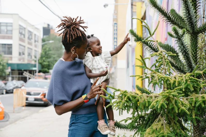 A mother holding her child points at a tree on a city street, showcasing urban nature and family bonding.