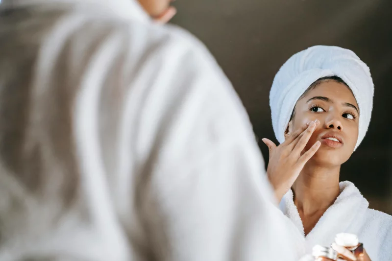 A young woman applies skincare cream while wearing a bathrobe in front of a mirror.